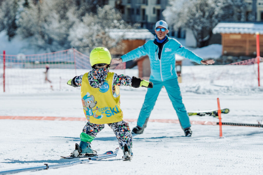 Photo prise de jour en hiver en format portrait. Nous pouvons voir le jardin d'enfants de l'ESI l'école de ski internationale. Nous pouvons voir un enfant en cours de ski accompagner d'un moniteur.