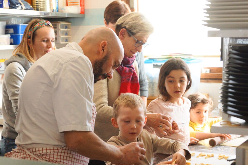 Chef cuisiner en train de montrer sa technique pour fabriquer des bugnes à des enfants durant la semaine de la gastronomie. La scène se passe dans la cuisine des Soldanelles