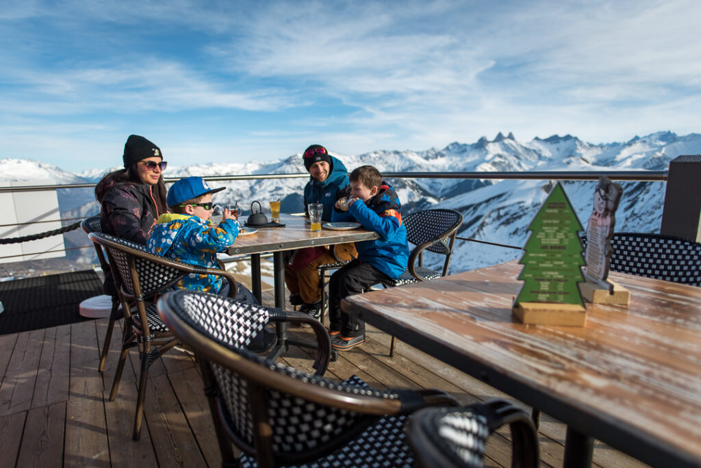 Photo prise en format paysage, de jour. Nous voyons une famille de deux parents et deux enfants en terrasse d'un restaurant d'altitude. Au premier plan il y a une table vide. Au second plan, les enfants sont en train de manger une crêpe. A l'arrière plan nous voyons le paysage avec notamment les Aiguilles d'Arves.