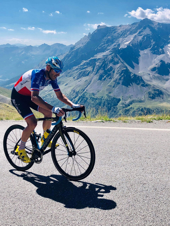 Portrait photo taken in summer. Williams Turnes on his bike, wearing his French Champion jersey, climbing the Galibier pass. William is in profile in the foreground, with mountains in the background.