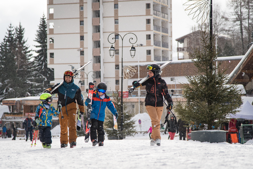 Photo prise au format horizontal où nous pouvons voir une famille en tenue de ski marchant sur de la neige avec les skis sur l'épaule pour aller en direction des remontées mécaniques