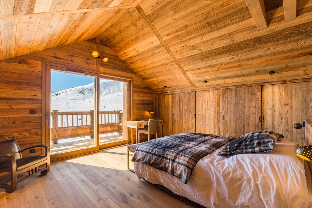 Vue sur la chambre d'un appartement en location à la résidence L'Odyssée de La Toussuire.. Lit king size, plafonds et parquets lambrissés, un petit bureau, un fauteuil en cuir vieilli confortable, un balcon et la magnifique vue sur le front de neige pour un appartement au pied des pistes.