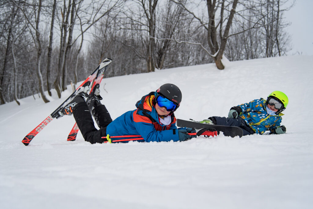 Photo au format horizontal en plein journée d'hiver. Nous pouvons voir deux enfants équipés en tenue de ski allongé au sol avec un grand sourire
