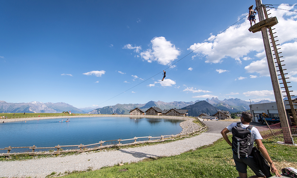 Photo prise de jour au lac de l'Eriscal. Nous pouvons voir une personne survolant le lac à l'aide de la tyrolienne. Le ciel bleu représente la moitié de l'image.