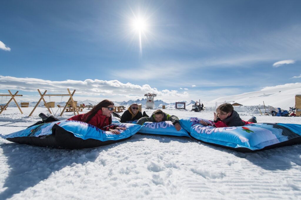 Photo taken in winter on a beautiful sunny day. A family of four lying on their stomachs on large cushions. They are enjoying the Snowpy Mountains fun zone.