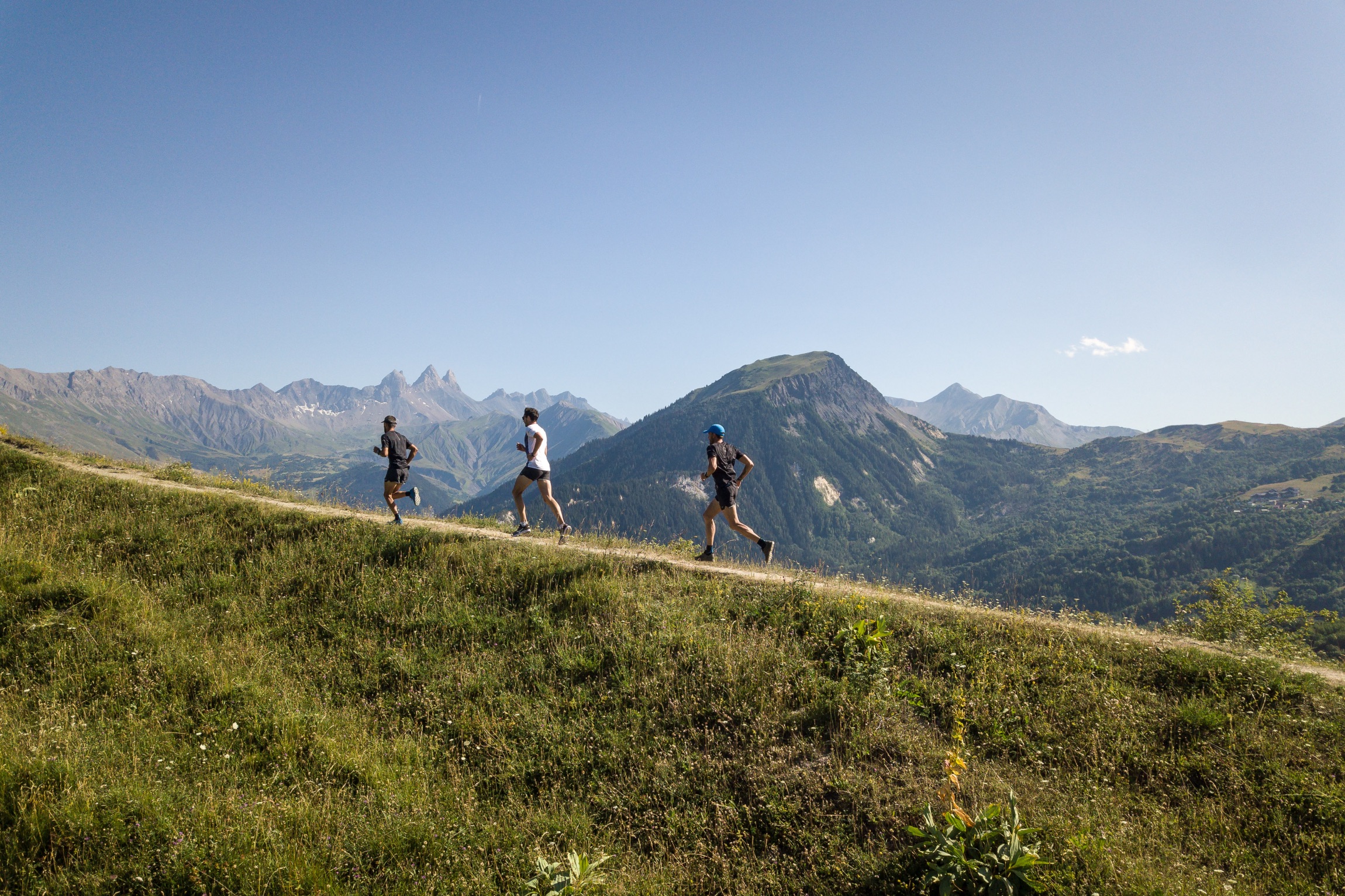 Trois personnes à la file indienne en train de courir sur un chemin de randonnée en montée. En fond nous pouvons apercevoir les Aiguilles d'Arves, le Mont Charvin ainsi que la Cime des Torches