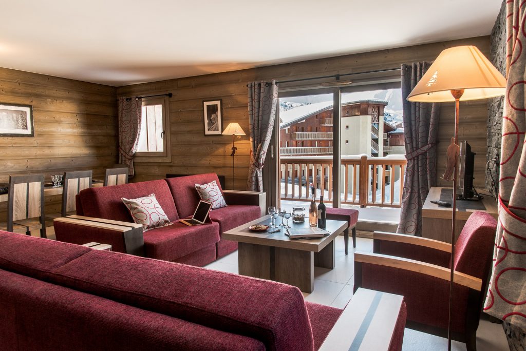 Vue sur salon et salle à manger d'un appartement de la résidence de tourisme l'Alpaga à La Toussuire, gérée par CGH. Des murs lambrissés de bois, des canapés roses moelleux et confortables, une grande table à manger. L'apéritif est servie sur la petite table du salon et la vue par la baie vitrée semble agréable. Bon séjour à La Toussuire!