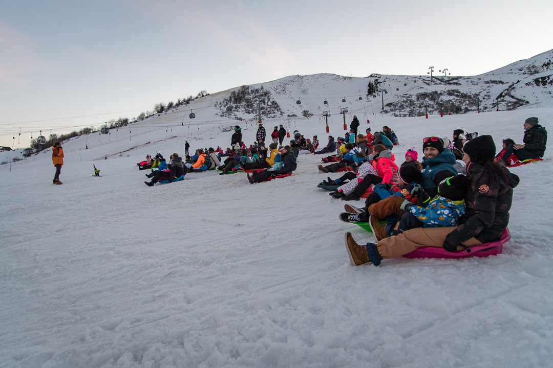 Photo prise en format paysage en fin de journée, où l'on voit de nombreuses personnes au départ d'une descente en luge géante sur les pistes de ski après la fermeture du domaine