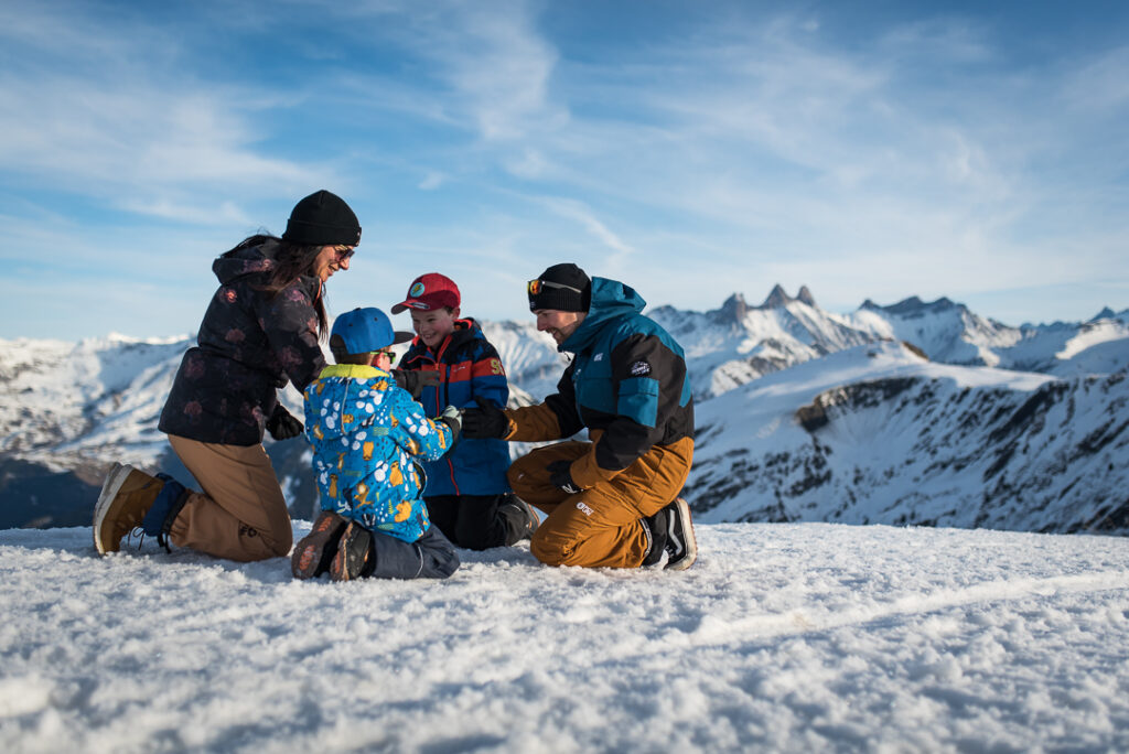 Photo prise de jour en format paysage. Au premier plan nous voyons une famille avec deux parents et deux enfants, qui sont à genoux dans la neige en train de jouer ensemble. A l'arrière plan, nous pouvons observer les montagnes avec les remarquables Aiguilles d'Arves.