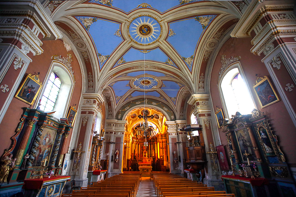 Foto van binnenkant van kerk van Fontcouverte. De foto is gecentreerd in de kerk met banken in het midden, schilderijen en vensters aan de zijkanten, het kruis met het hotel op de achtergrond en het blauwe plafond erboven