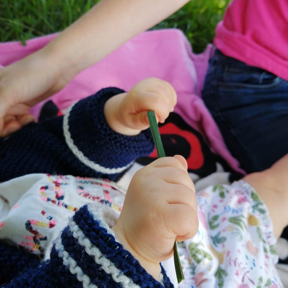 Un bébé couché sur une couverture sur l'herbe prend le frais à l'ombre d'un arbre, accompagné d'une nounou de la crèche, halte garderie du Pôle Enfance La Ruche de La Toussuire