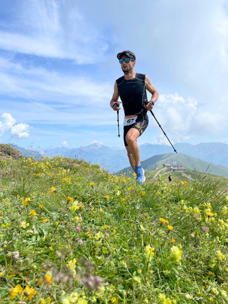 Traileur équipé de bâton de marche est en train de finir le demi kilomètre vertical de la Toussuire. La photo est prise en contre plongée où l'on peut voir la flore des montagne et les paysage au loin.