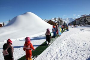 In one of the two La Toussuire skiing schools early-learning areas, children line up on a mat, waiting for an instructor to give them skiing instructions. A snow-covered mountain is in the background.