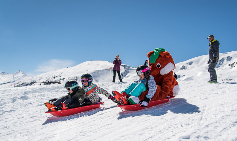 Siblings engage in a sledding competition, under their parents' gaze. Toussy, La Toussuire's mascot, takes part in the race in a sled.