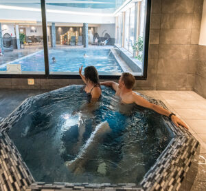 Photo in a spa. We see a couple in a jacuzzi in the foreground, gazing towards the background at their children, who are in a swimming pool.