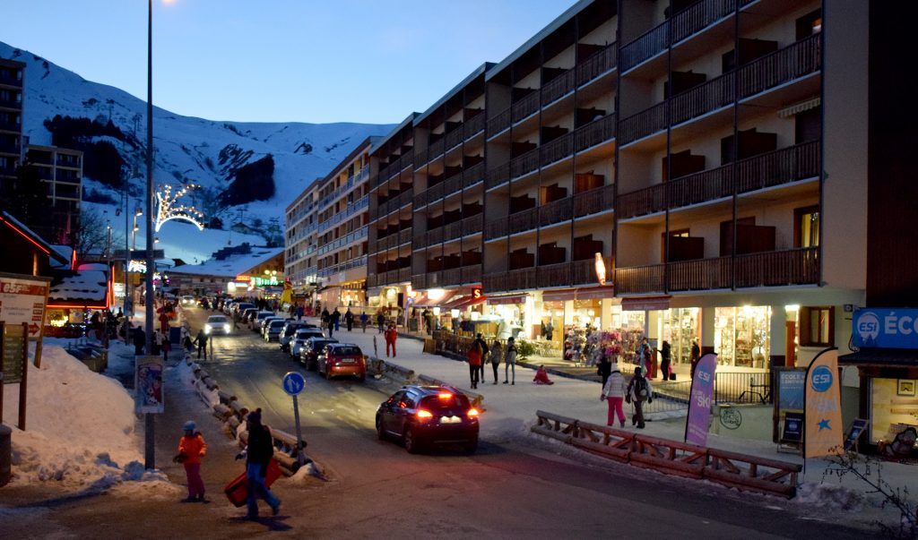 Photo prise en fin de journée d'hiver dans la rue verte. Nous pouvons voir des voitures sur la routes, des piétons sur la rue verte enneigés et les commerces ouvertes sous les arcades