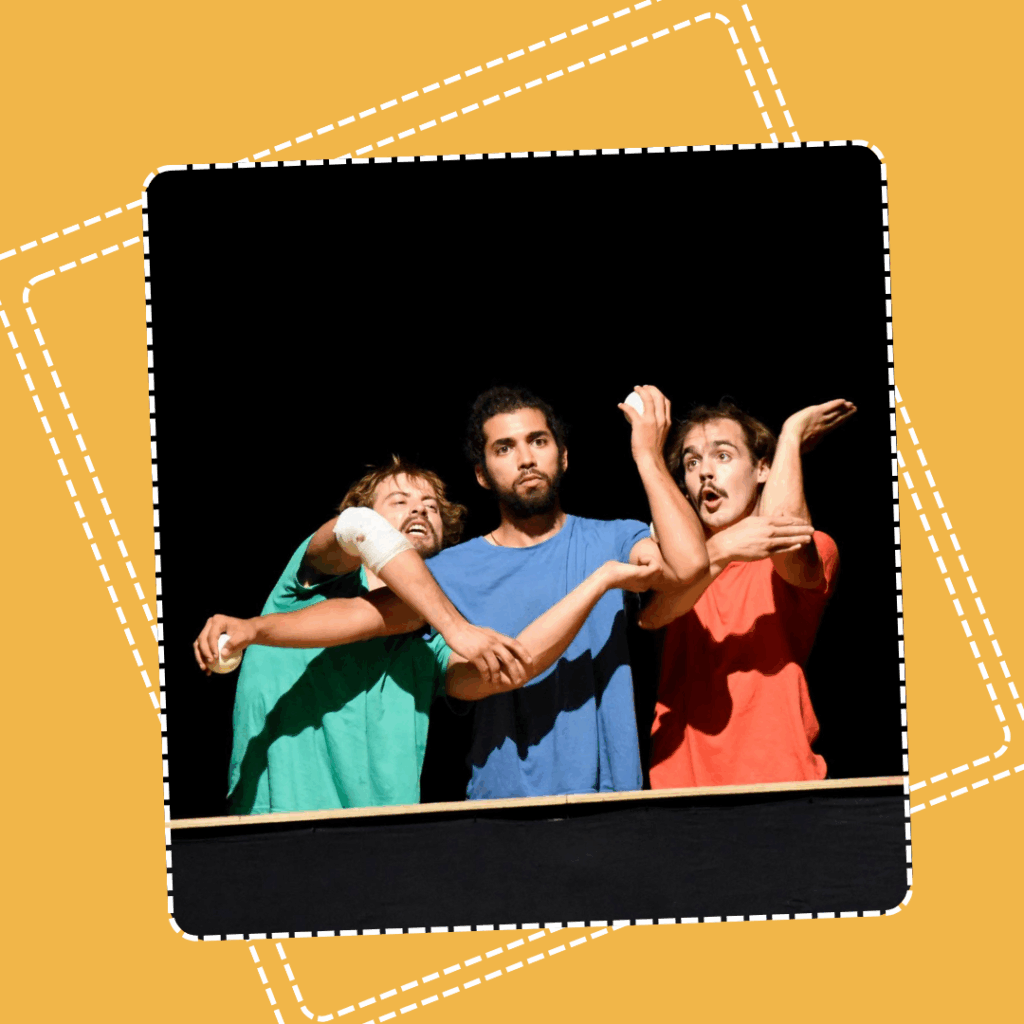 Trois comédiens vêtus de t-shirts colorés (vert, bleu et rouge) se tiennent serrés côte à côte sur scène, formant une composition burlesque avec leurs bras entremêlés. Leurs expressions faciales exagérées évoquent la surprise, la confusion ou l’amusement. L’arrière-plan noir met en valeur la gestuelle théâtrale et le jeu de scène.