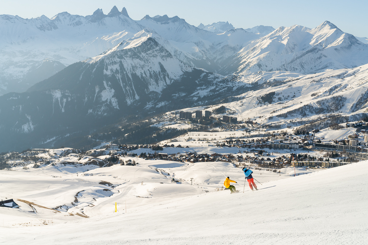 Daytime landscape shot in winter. We can see two people, one on skis, the other on a snowboard, descending a slope to reach the resort of La Toussuire, which we can see below. 